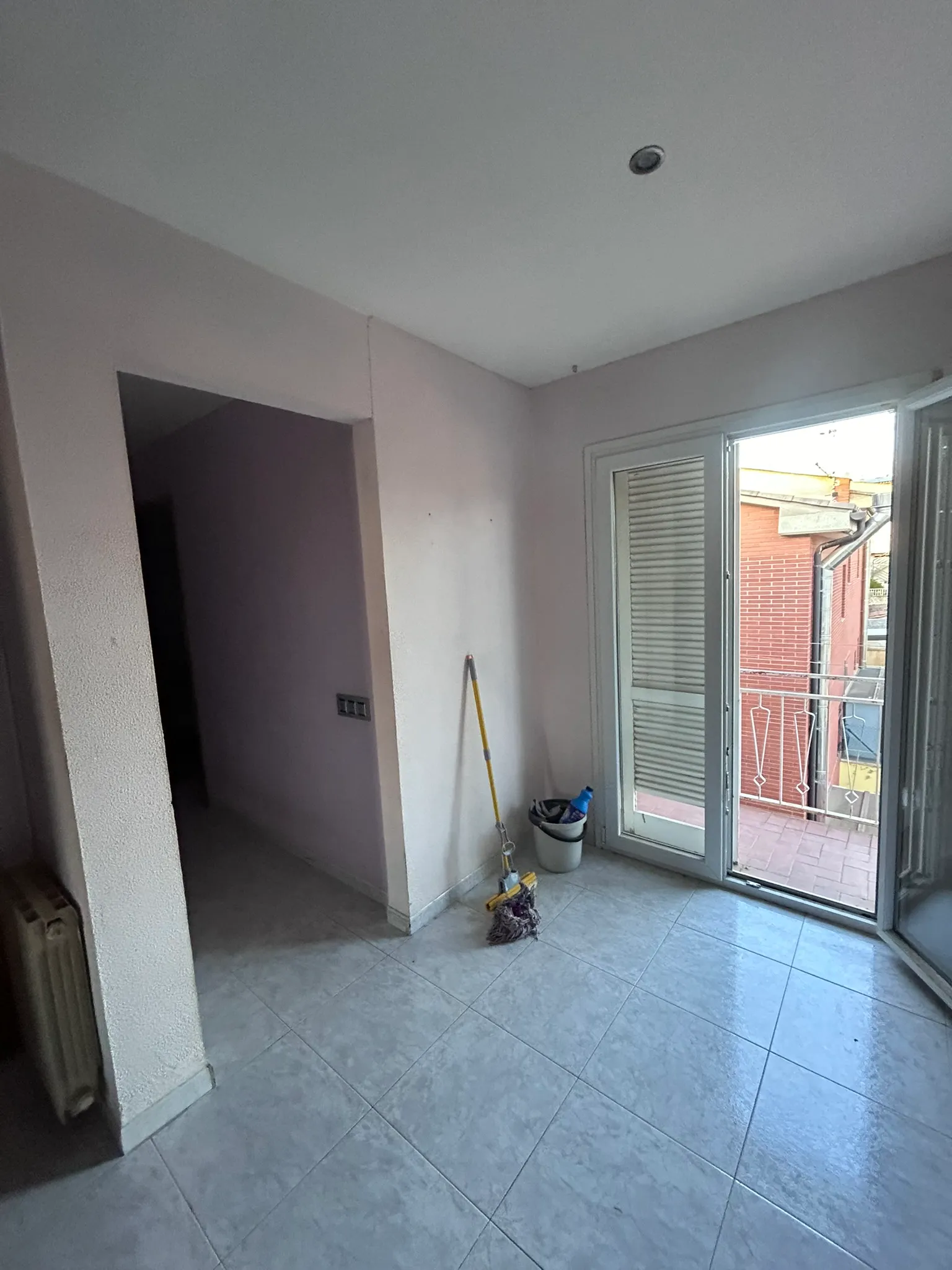 Estancia de la vivienda antes de la reforma: paredes con gotelé, suelo de baldosa gris, radiador antiguo, puerta de paso a otra habitación y balcón de aluminio blanco