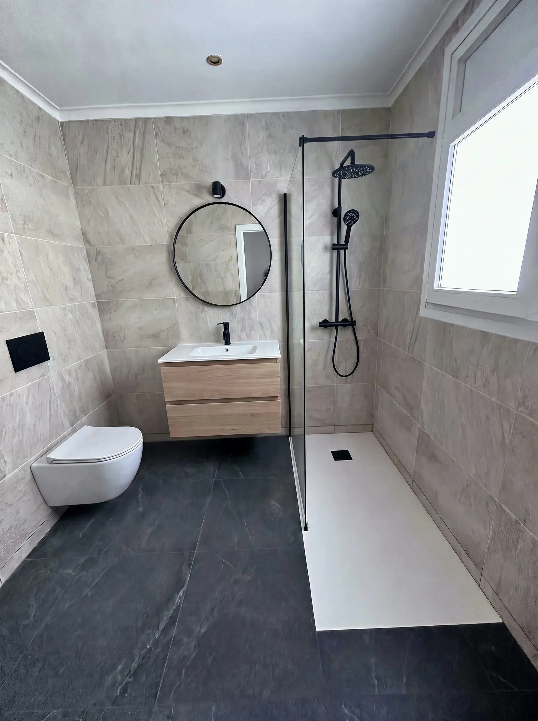 Baño reformado en Banyoles: columna de ducha negra, mampara fija, inodoro suspendido y mueble de roble sobre porcelánico efecto piedra