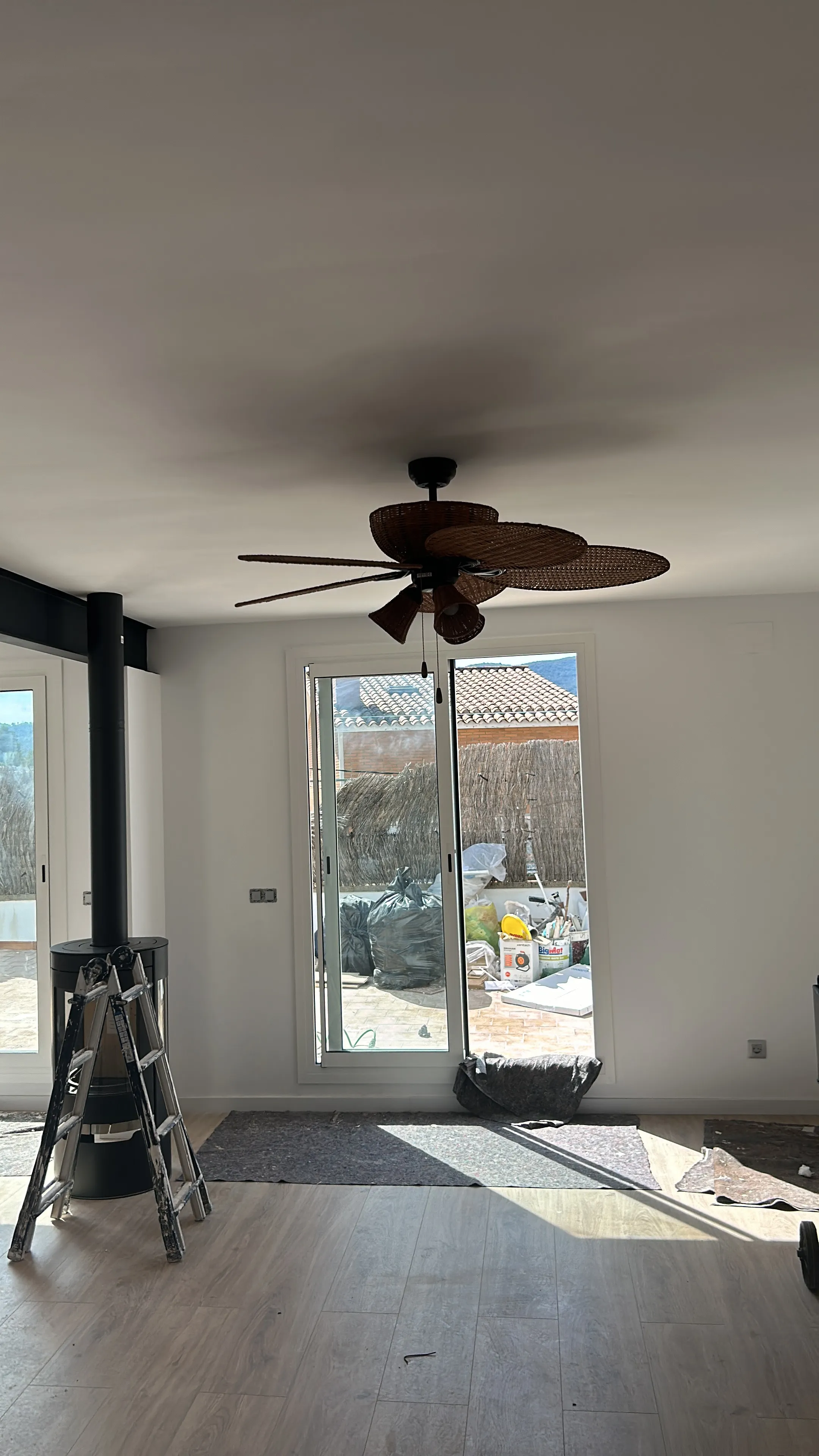 Salón pintado en blanco con ventilador de techo y ventanales al patio; en un extremo del techo asoma el arranque de la viga vista negra y, en primer plano, el tubo negro de la estufa de leña sube hasta el techo, con una escalera de tijera y mantas de protección todavía en la estancia