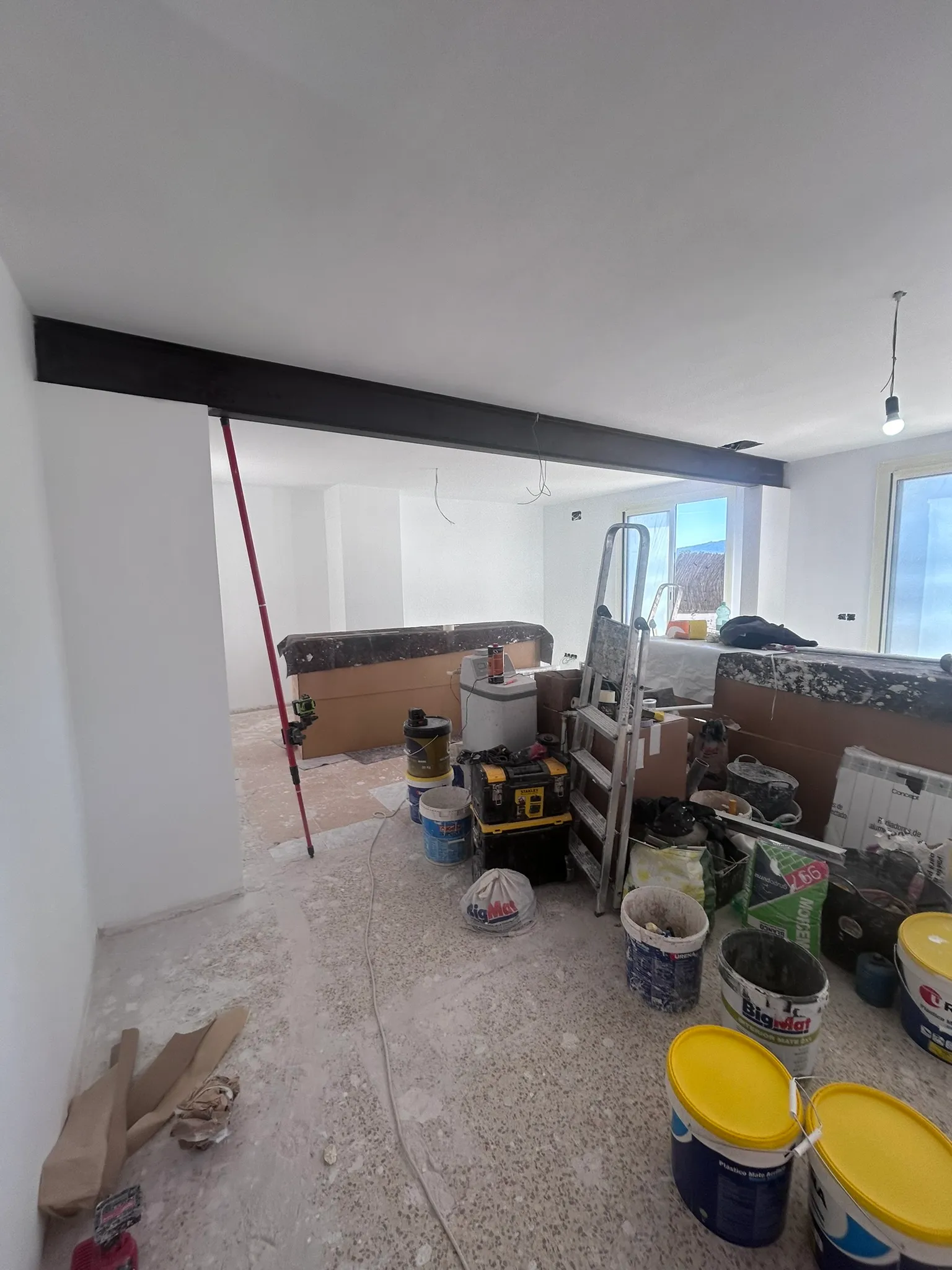 Viga vista pintada de negro cruzando de lado a lado el hueco abierto entre dos estancias; a través del vano se ve la siguiente sala con el suelo aún sin acabar