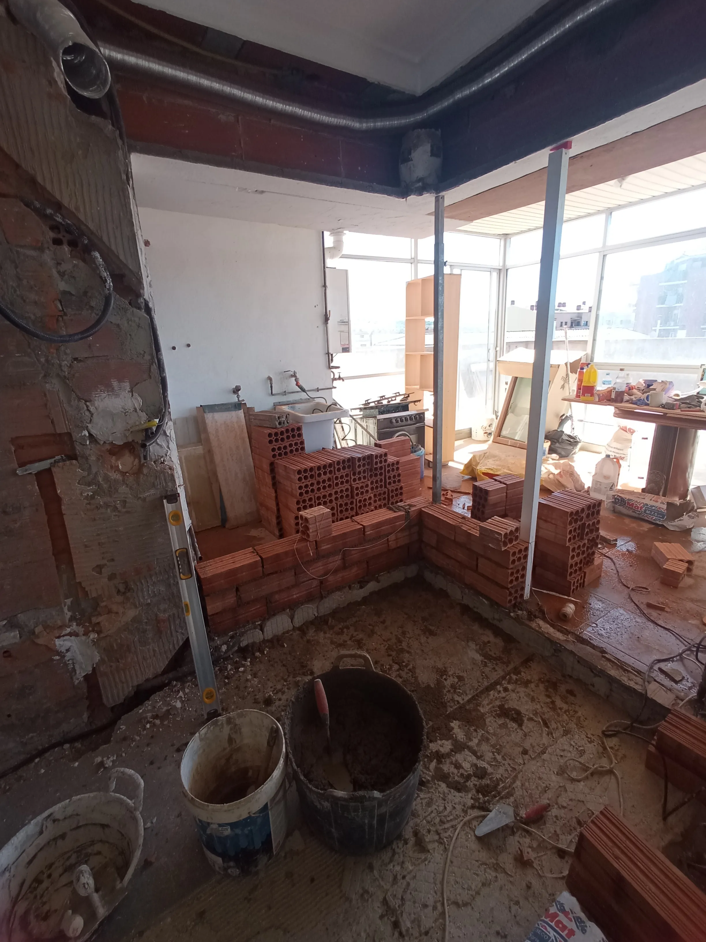 Obra en curso: tabique de ladrillo hueco recién levantado, puntales metálicos apoyados contra el techo, ladrillos apilados, capazos de mortero y un nivel de burbuja apoyado contra la pared, sobre el pavimento existente aún sin retirar