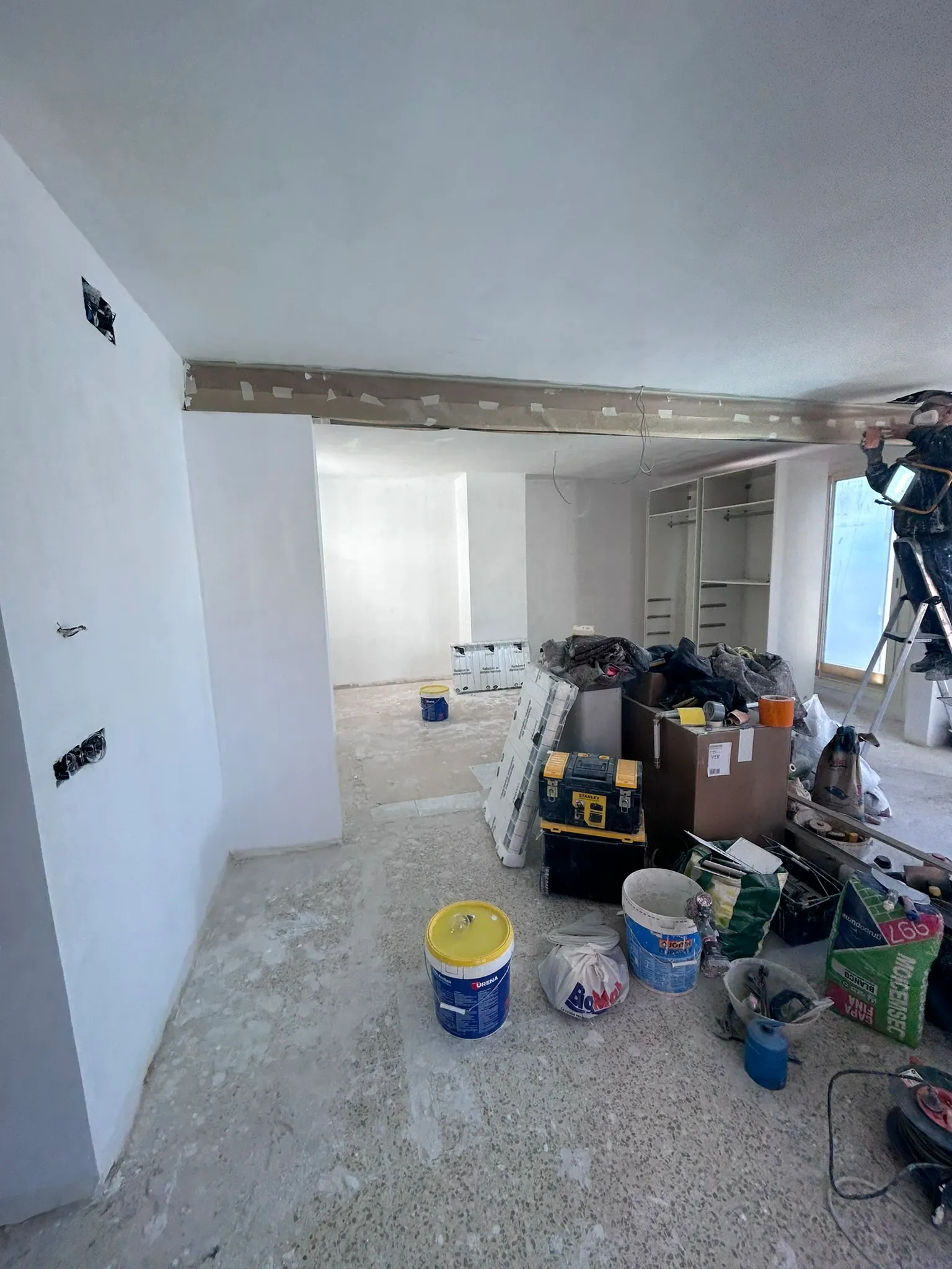 Operario subido a una escalera de tijera rematando el encuentro del techo, con los armarios empotrados ya montados al fondo y sacos de mortero y cubos de pintura en primer plano