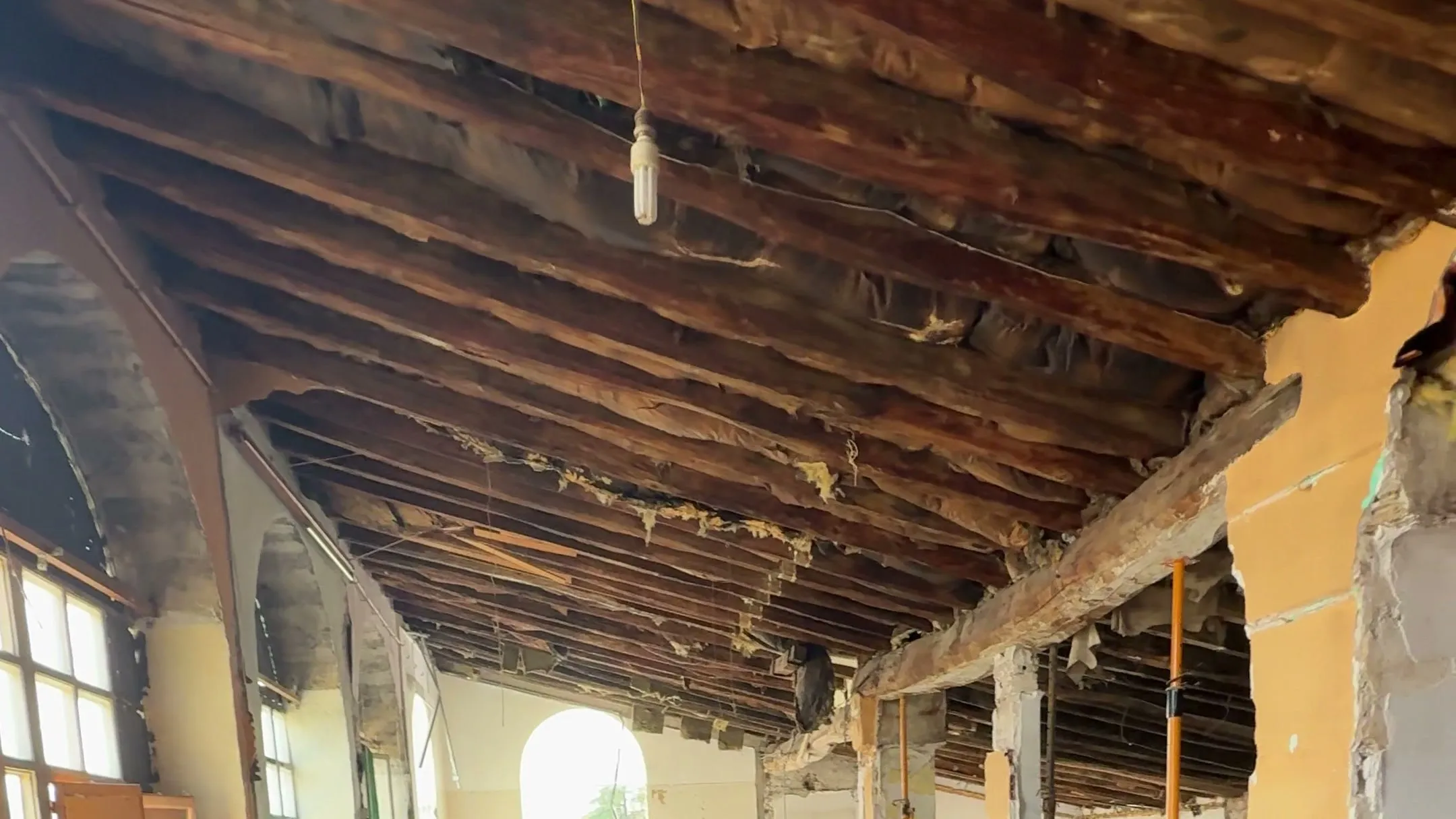Interior de un edificio en rehabilitación en Banyoles: techo de vigas de madera al descubierto y ventana de arco al fondo.